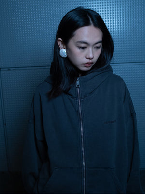 A person wearing a dark Carhartt WIP hoodie stands against a textured wall, looking down with a contemplative expression.