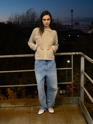 A person stands on a balcony at dusk wearing a beige Carhartt WIP sweater and blue jeans.